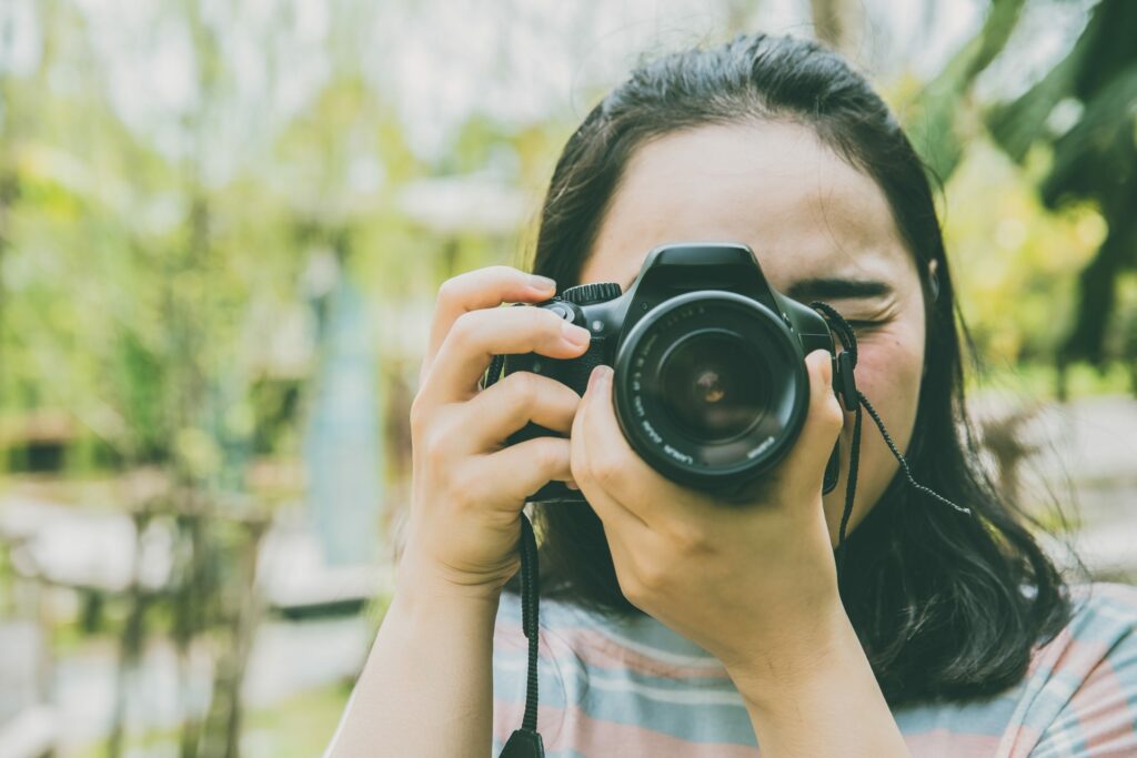 Individual holding a camera and taking a photo in an outdoor setting with greenery in the background.