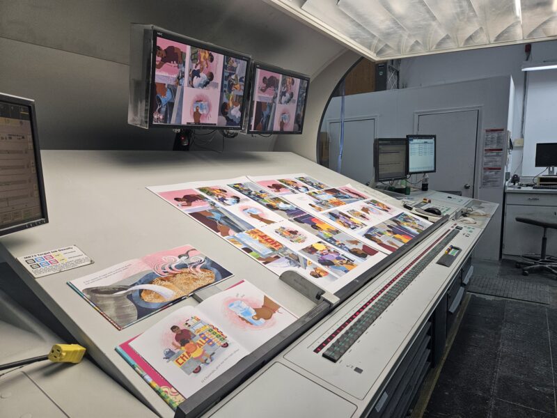 Illustrated print sheets laid out on a proofing station beneath inspection lights and monitor displays.