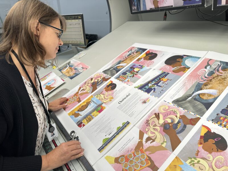 Person reviewing large illustrated print sheets at a proofing station under bright inspection lighting.