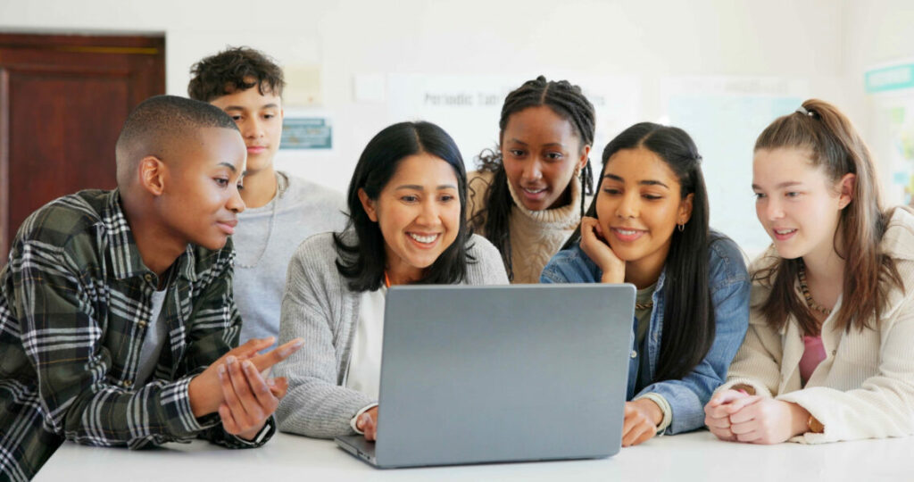 Group of students gathered around a laptop, working together in a bright classroom setting.