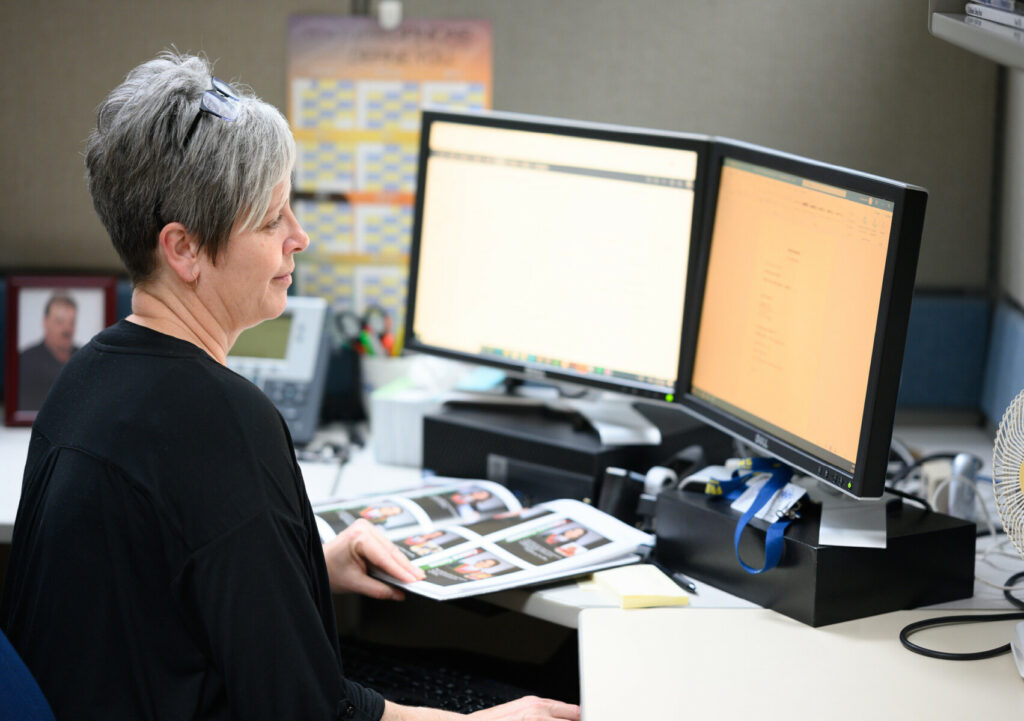 Employee working at a dual‑monitor workstation while reviewing printed photo pages.