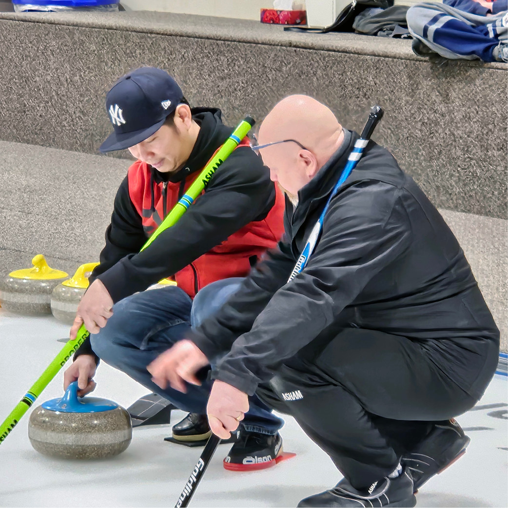 Two people kneeling on the ice while preparing to deliver a curling stone with their brooms.