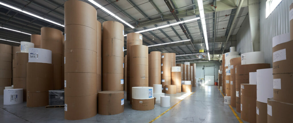 Large rolls of paper stored in rows inside a spacious book printing facility.
