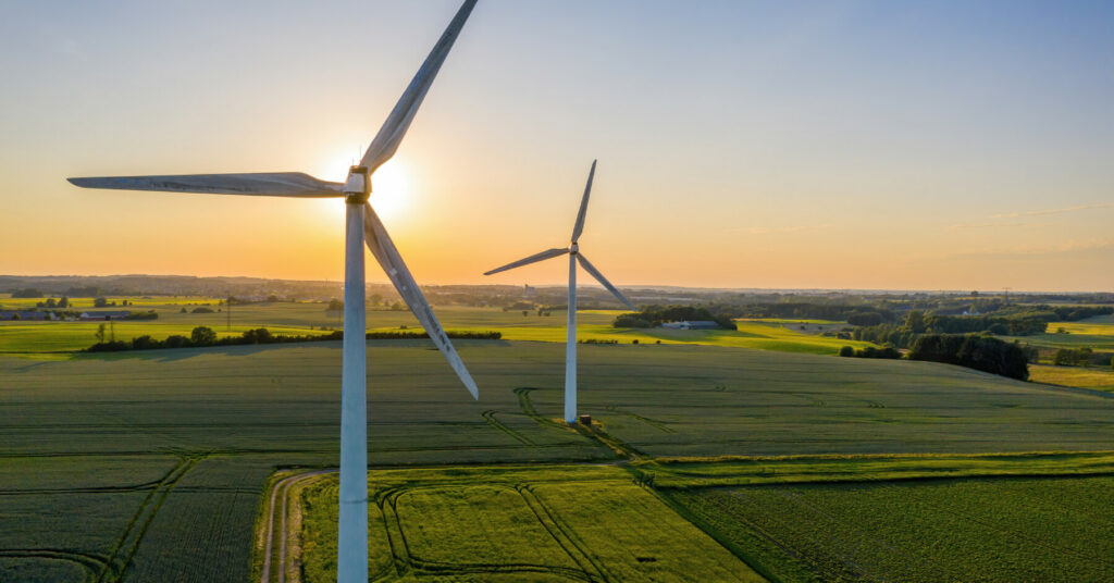 Two wind turbines standing in a green field with the sun setting behind them.