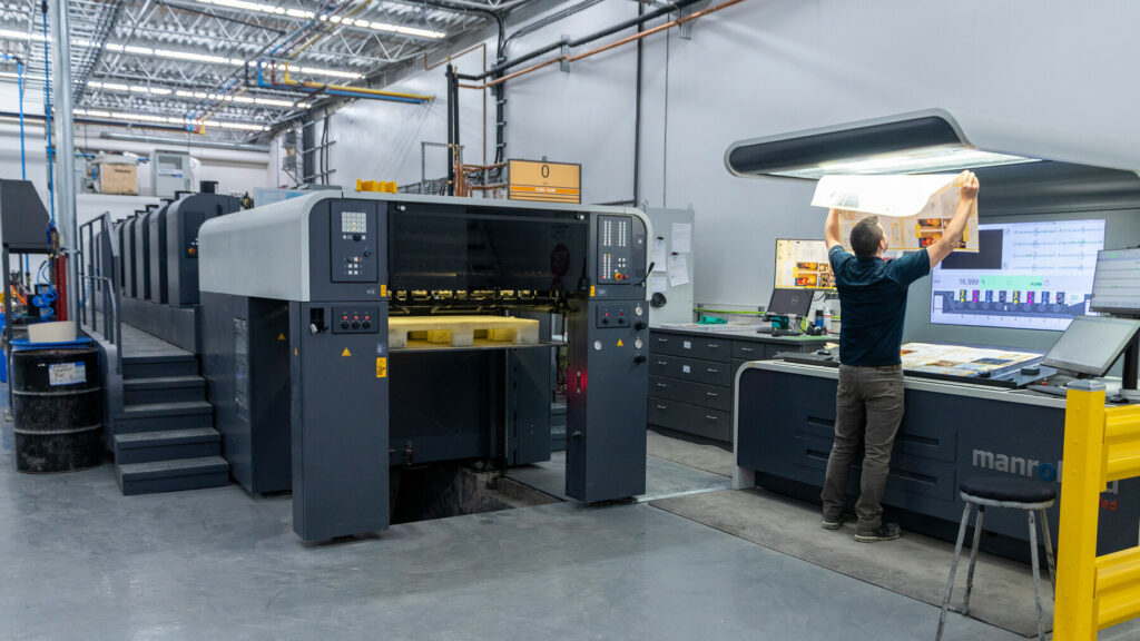 Operator reviewing a printed sheet under inspection lights beside a large offset press in a production area.