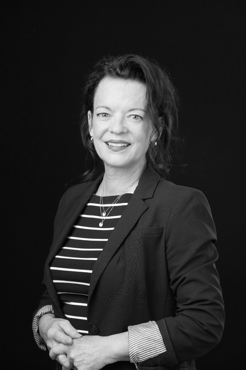 Person wearing a blazer and striped shirt standing against a dark background with hands lightly clasped.