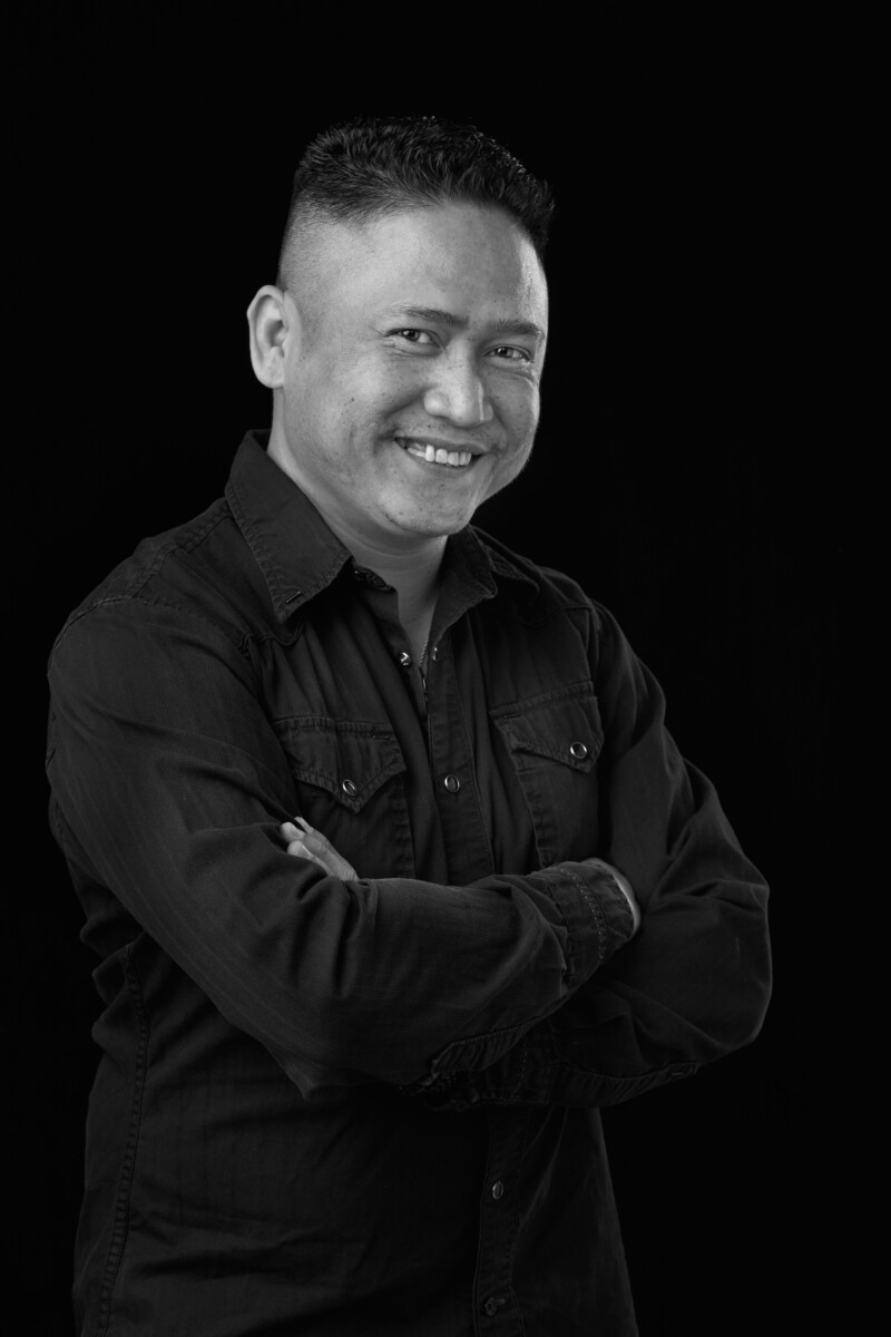 Person standing with arms crossed, wearing a dark button‑up shirt against a black background.