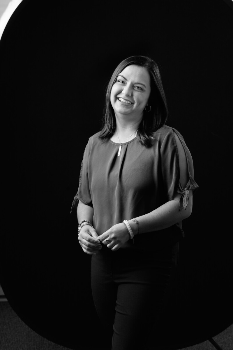 Person standing in front of a dark backdrop wearing a short‑sleeved blouse with hands loosely clasped.