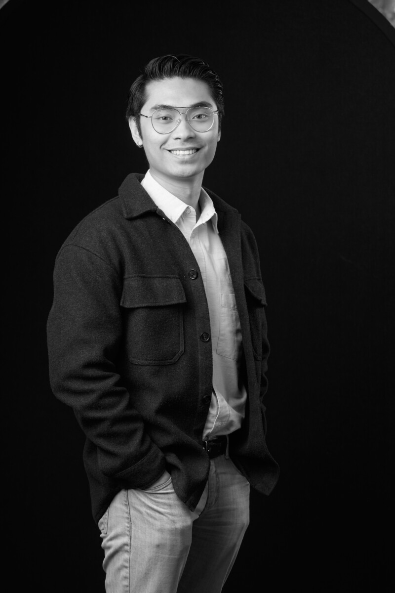 Person standing with one hand in a pocket, wearing a button‑up shirt and jacket against a dark backdrop.