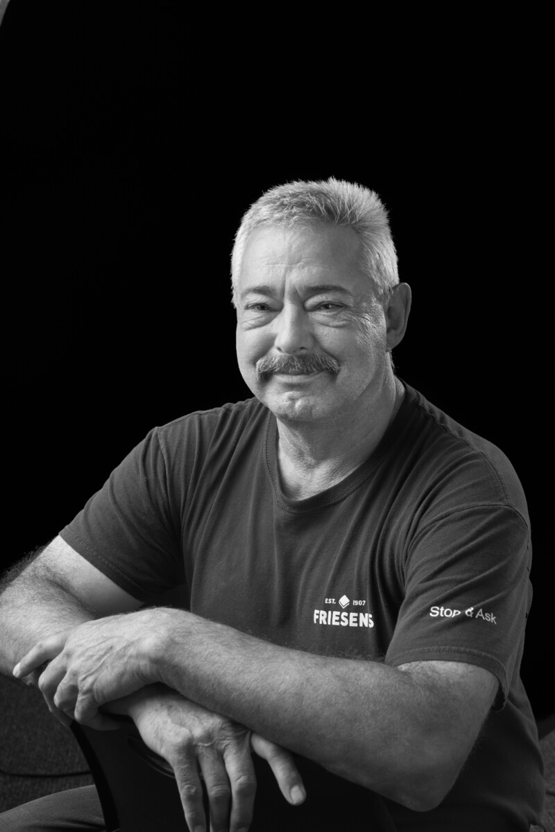 Person seated against a dark background wearing a Friesens T‑shirt with arms resting on a chair.
