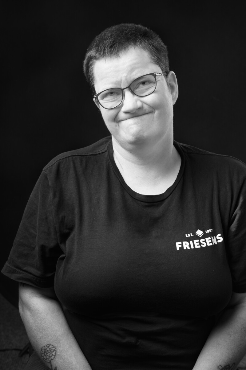 Person seated against a dark background wearing a Friesens T‑shirt with arms relaxed at their sides.