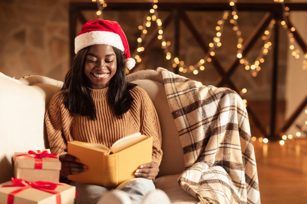 Person in a Santa hat reading a book on a sofa with gifts and warm holiday lights in the background.