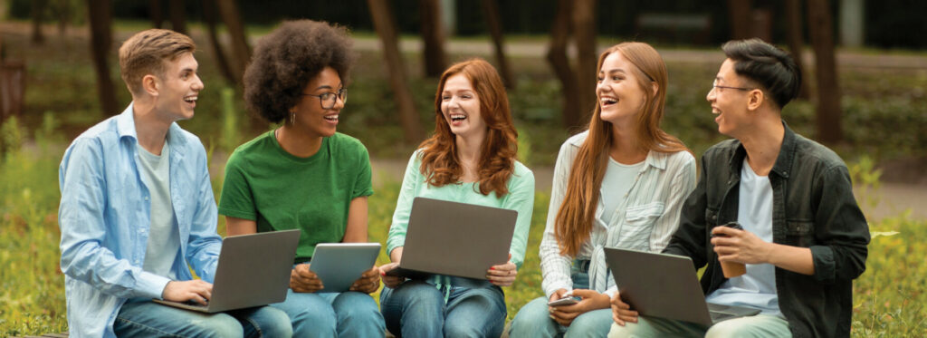Group of students sitting together outside using laptops and tablets while talking and studying.