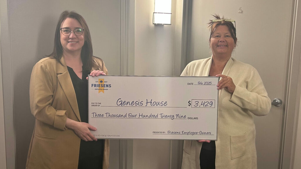 Two people holding a large donation cheque for Genesis House inside a hallway.