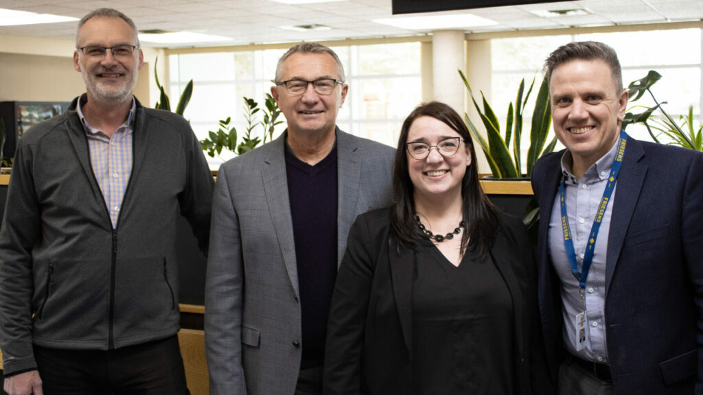 Four employees standing together in an office area with plants and natural light in the background.