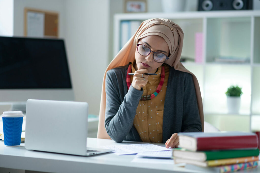Person working at a desk with a laptop, documents, books, and a cup of coffee in a bright office.