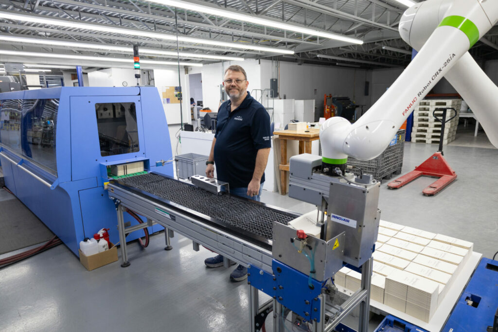 Person working beside an inkjet press and conveyor system with a robotic arm in a print production area