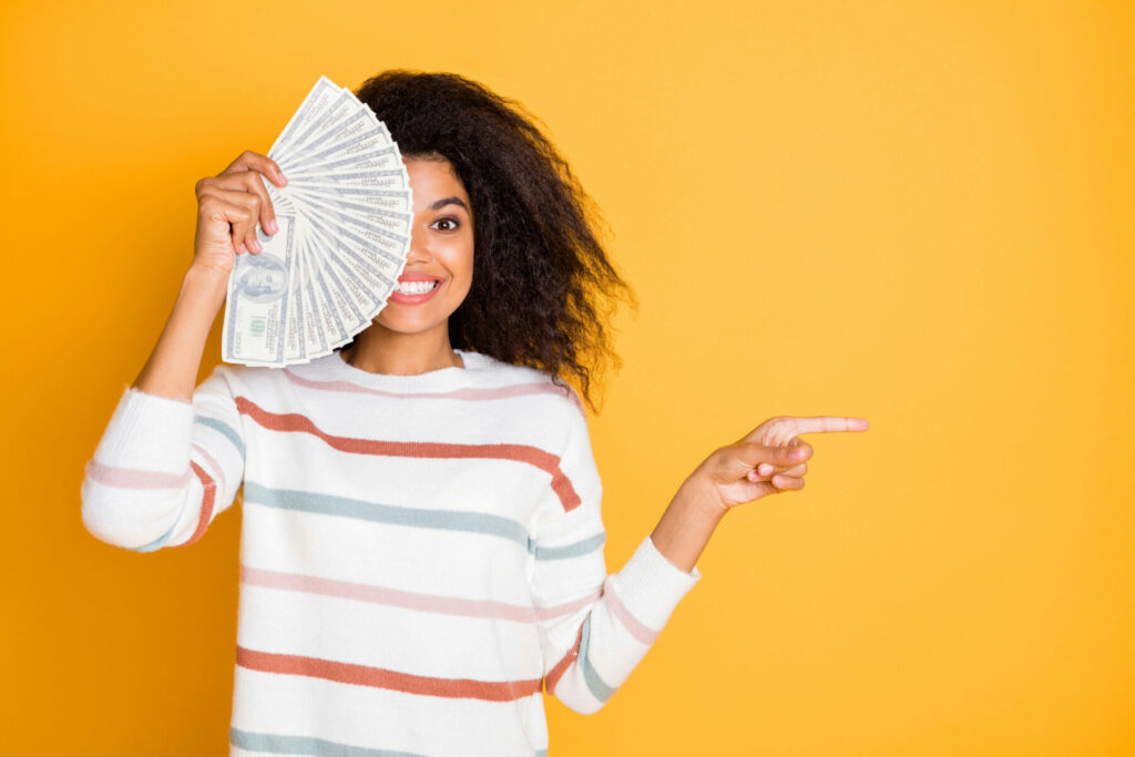 Person holding a fan of cash in one hand while pointing to the side against a bright yellow background.
