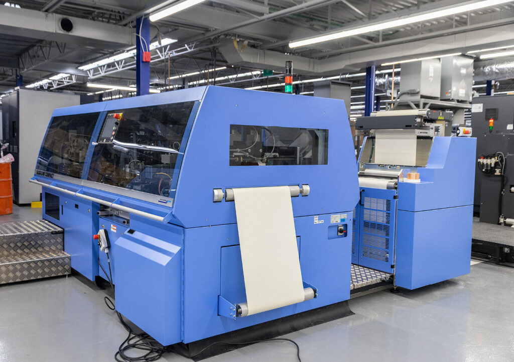 Blue industrial inkjet press unit with paper feed inside a production facility.