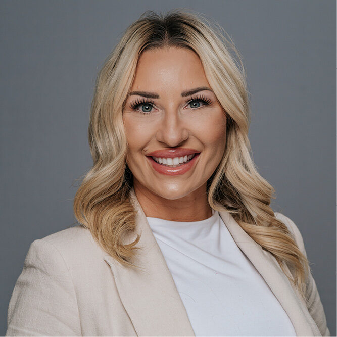 Person wearing a light blazer and white top posing against a plain gray background.