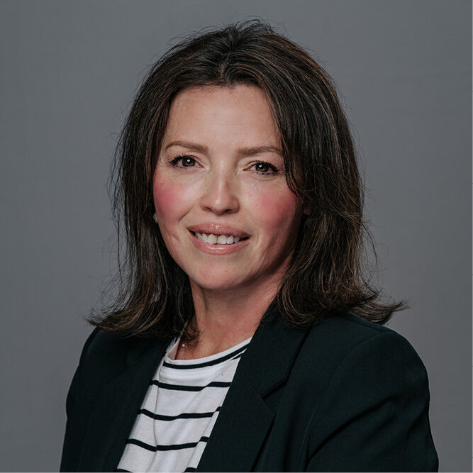 Person wearing a dark blazer and striped shirt against a plain grey studio background.