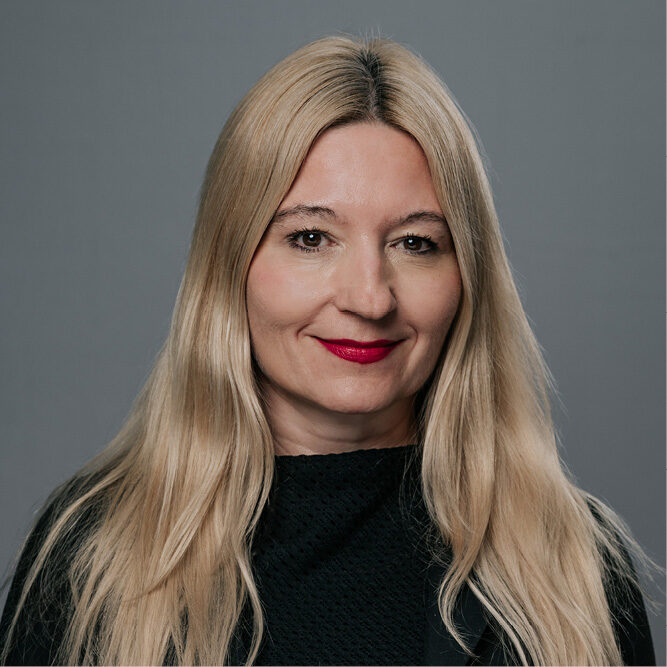 Person with long blonde hair wearing a black top, posed against a plain gray background.