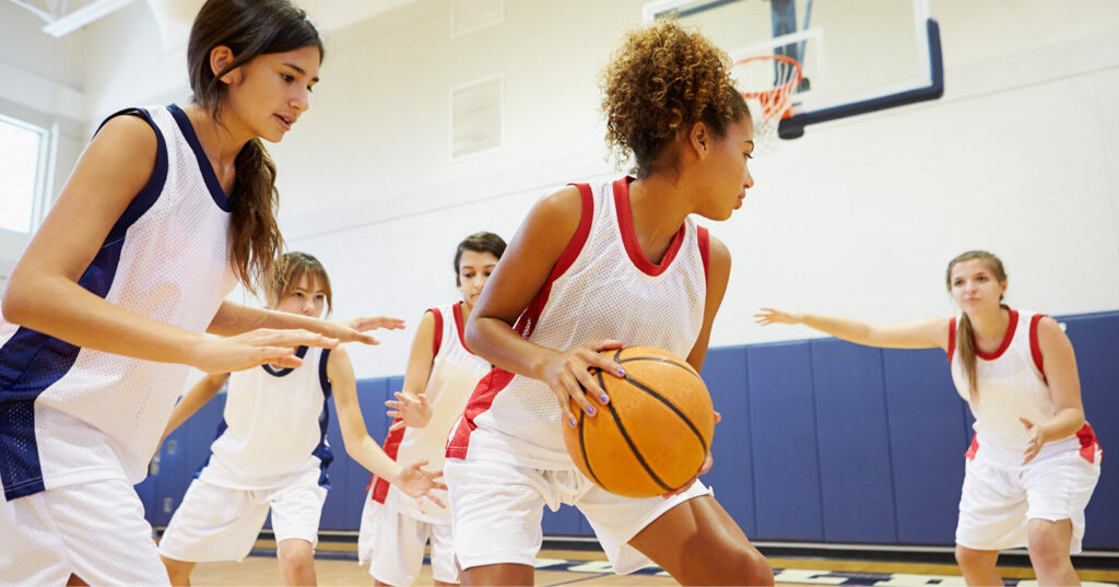Players in a girls’ basketball game defend as a teammate holds the ball on the court.