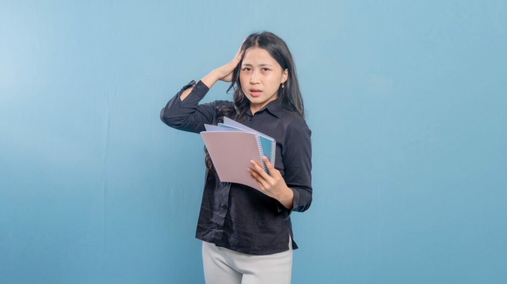 Person standing against a blue background holding several notebooks with one hand on their head.