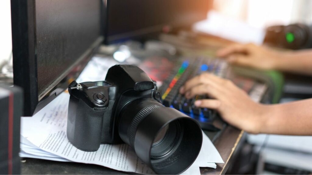 Digital camera resting on papers next to a computer monitor while someone types on a keyboard.