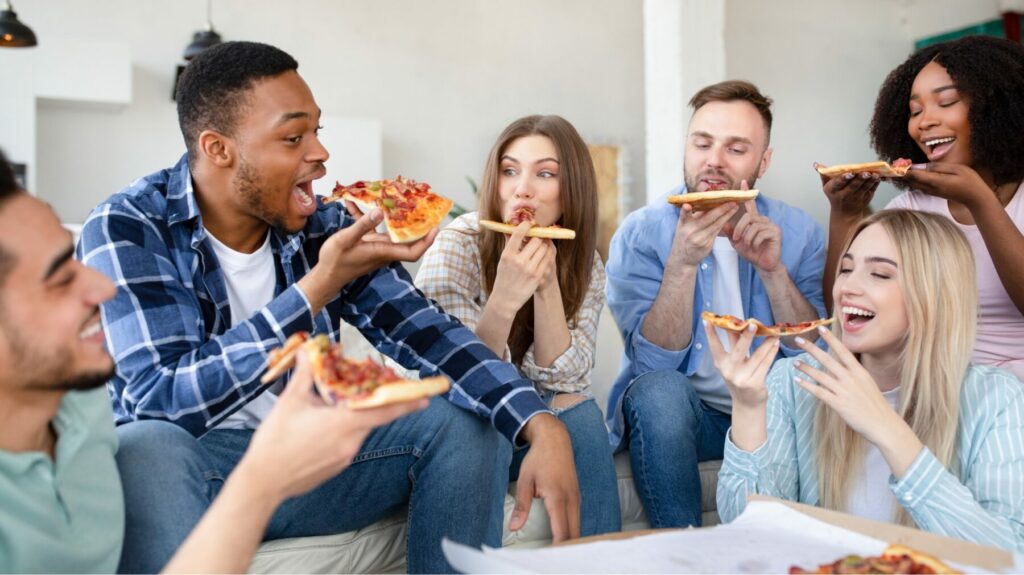 People sitting together indoors, sharing and eating slices of pizza in a casual gathering.