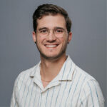 Person wearing a light striped shirt posing for a studio headshot against a grey backdrop.