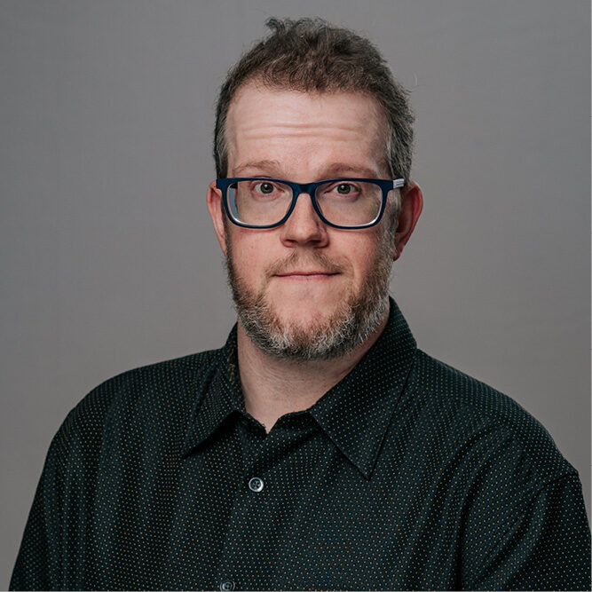 Man wearing a dark patterned button‑up shirt against a neutral gray background.
