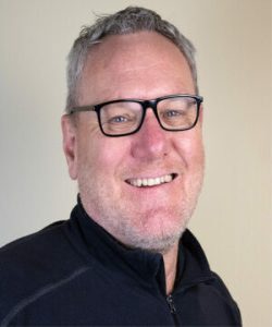 Person wearing a dark zip‑up sweater standing against a plain light-colored background.