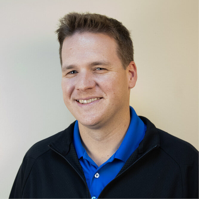Person wearing a blue polo and black jacket standing against a plain light-colored wall.