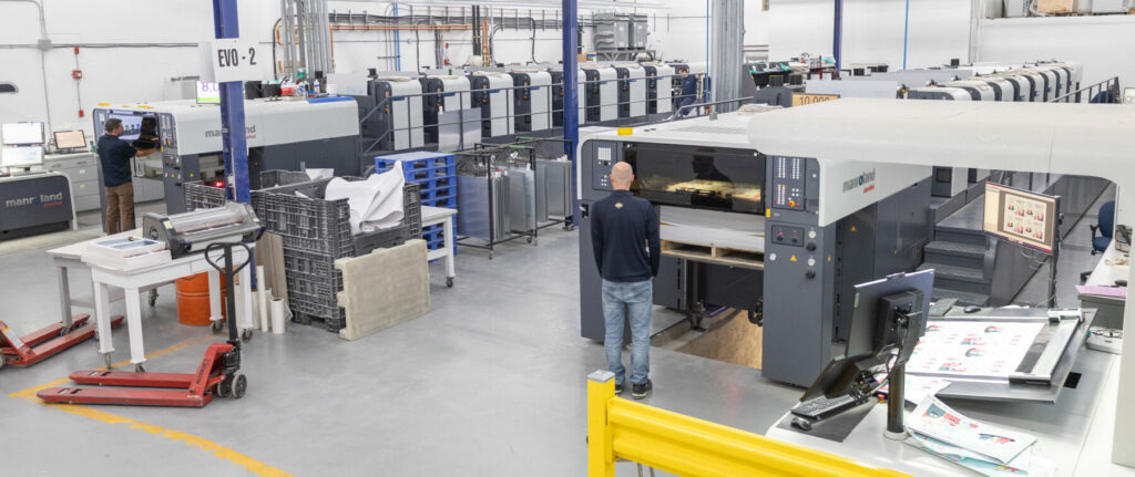 Large offset printing presses operating in a production facility with printed sheets and equipment visible.