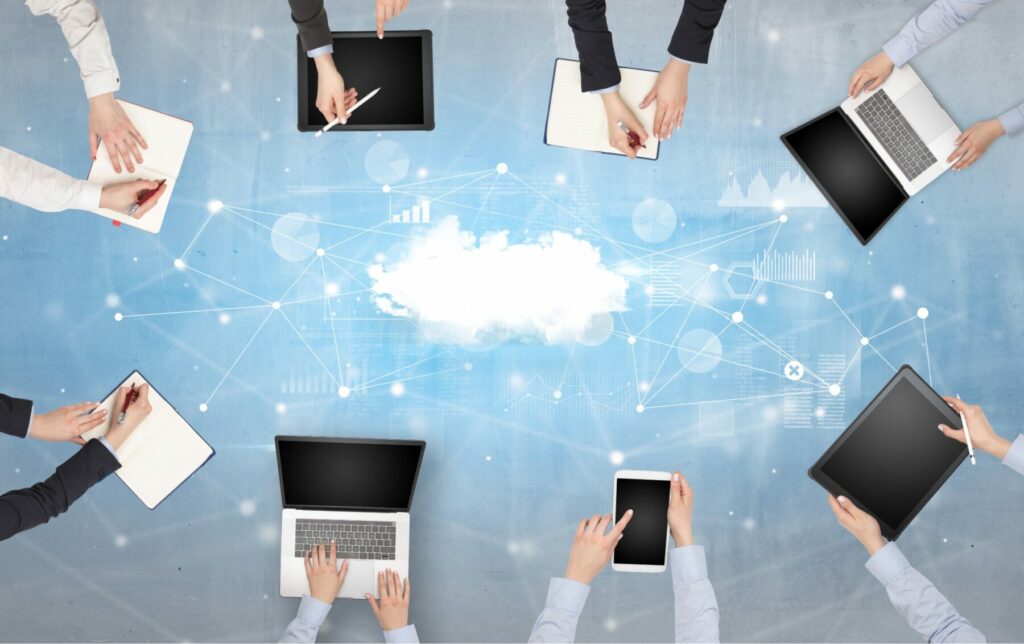 Multiple hands using laptops and tablets around a cloud graphic representing digital collaboration.