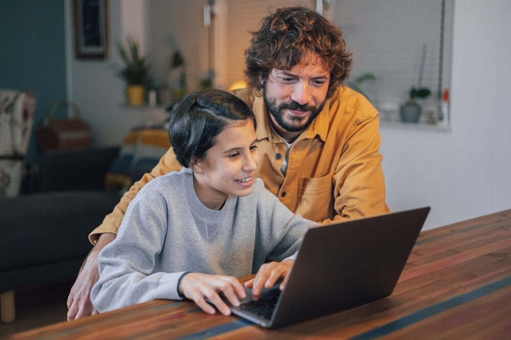 An adult standing beside a child seated at a table, both focused on a laptop in a home setting.