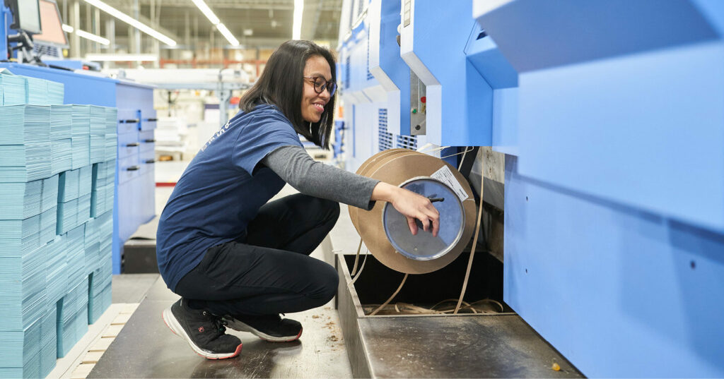 Worker replacing a roll of book headband material on a blue machine in a book manufacturing facility.