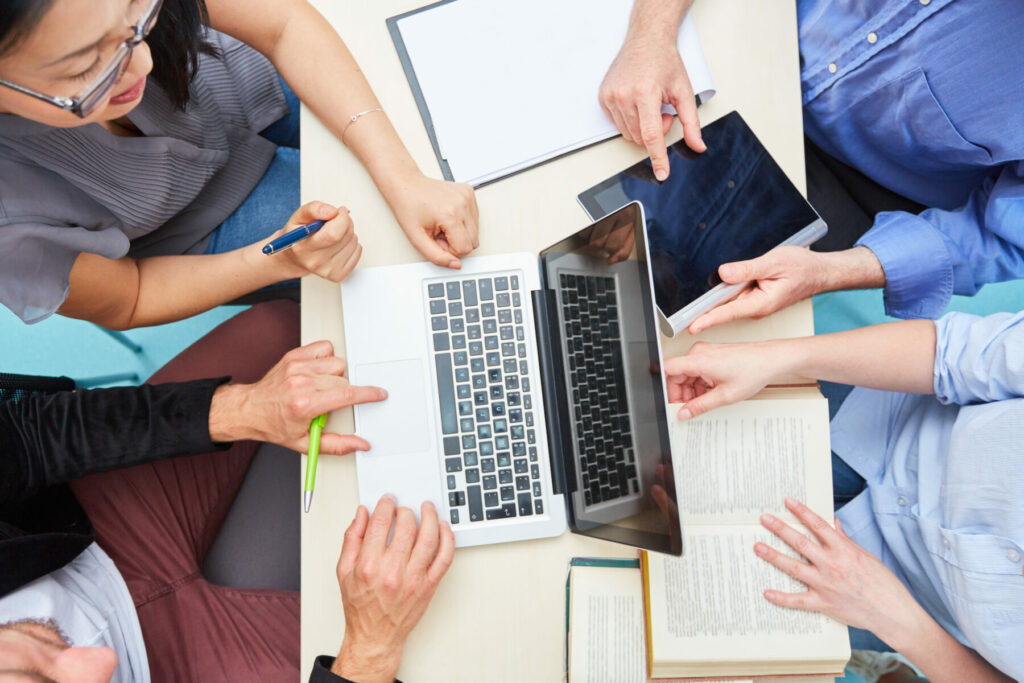 Top-down view of several people gathered around a table working with a laptop, tablet, notebooks, and documents.