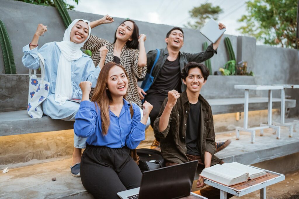 A group of people gathered outside, raising their fists in a celebratory pose beside benches, a laptop, and an open book.