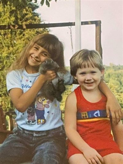 Two children sitting together outside, one holding a small pet while they pose on a wooden structure.