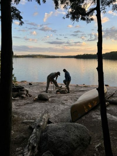 Two people preparing firewood on a rocky lakeside campsite at sunset with a canoe nearby.