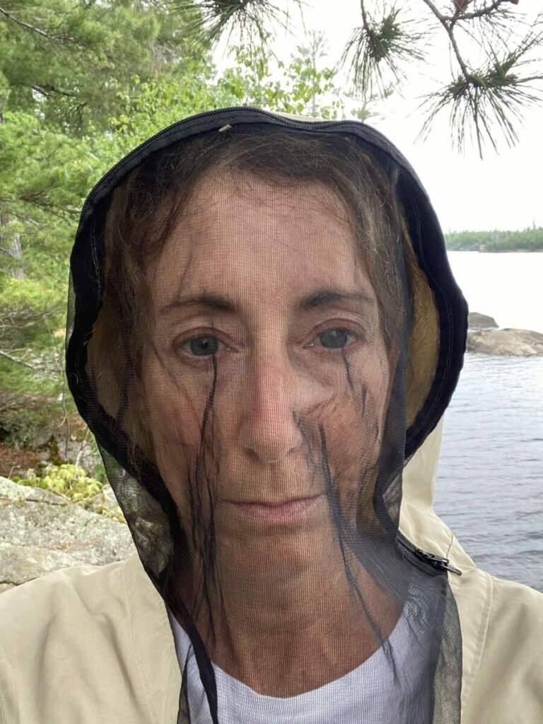 Person wearing a bug jacket near a rocky lakeshore with trees and calm water in the background.