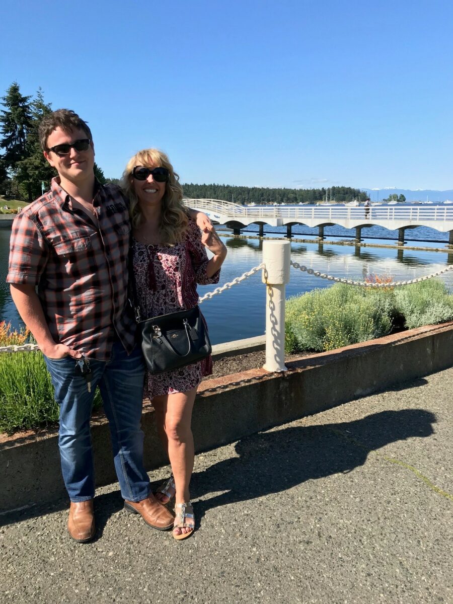 Two people standing by a calm waterfront with a white arched boardwalk bridge and trees under a clear blue sky.