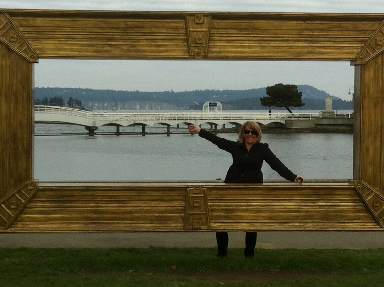 Person posing inside a large outdoor picture frame with a white arched bridge and calm water in the background.