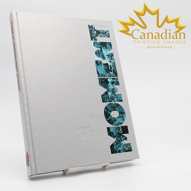 A white hardcover yearbook titled In the Moment, displayed upright with turquoise photo‑collage lettering, alongside a gold Canadian Printing Awards 2025 Winner emblem.
