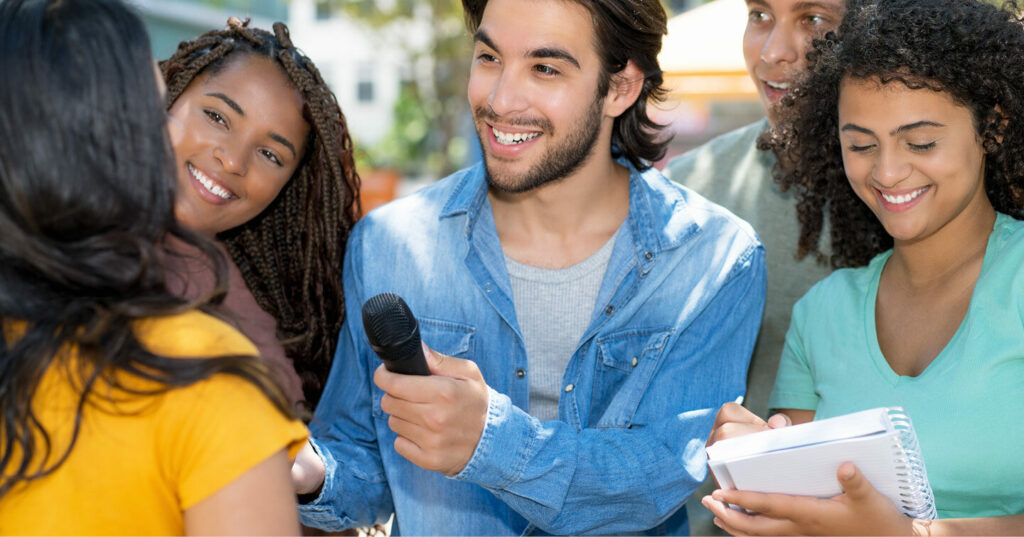 Several people gathered outdoors as one person holds a microphone and another takes notes during an informal interview.