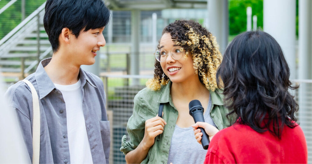 A person holding a microphone interviews two individuals standing outside near a staircase and metal railing in a bright, open area.