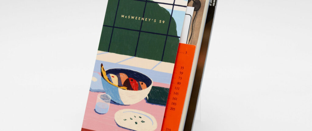 Angled view of a McSweeney’s 59 book featuring an illustrated stepback cover with a bowl of fruit and colorful layered inserts visible behind the front panel.