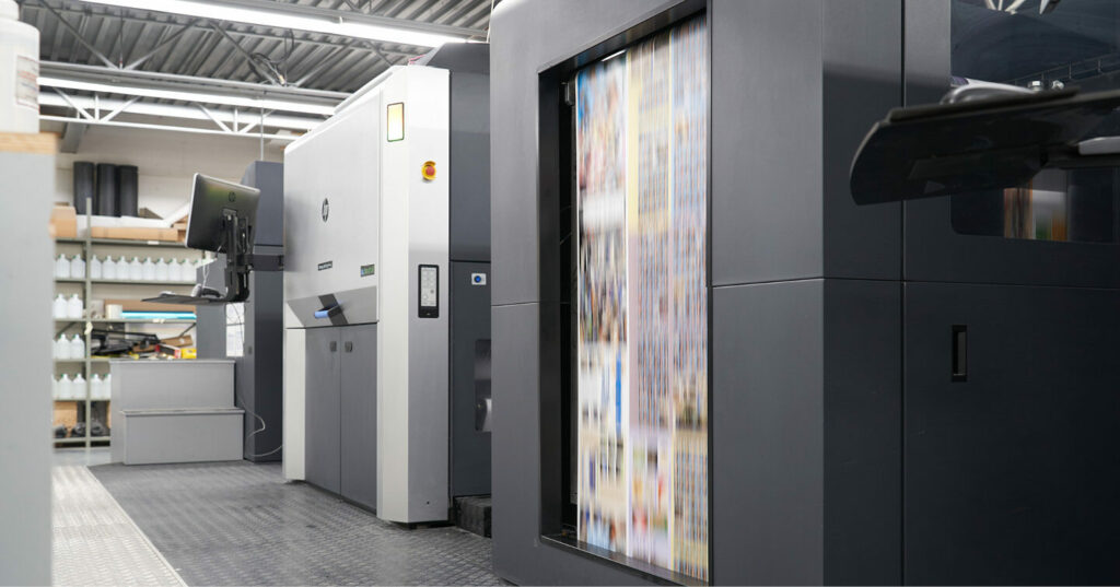 Digital toner press operating in a print facility, with a colorful sheet moving through the press and surrounding equipment visible.